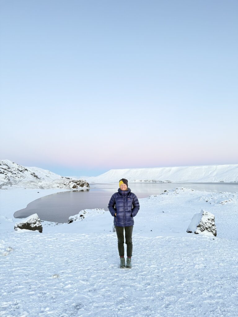 Kelionė į Islandiją žiemą. Moteris su pūkine striuke stovinti ant snieguoto šlaito su rožiniu saulėlydžio dangumi fone